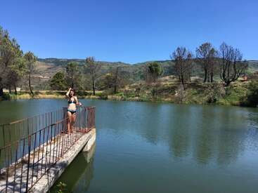 A woman in a swimsuit stands on a small bridge over a peaceful pond, surrounded by trees and hills under a clear blue sky.