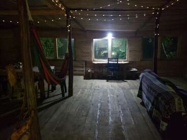 O interior de uma cabana de madeira aconchegante à noite, com luzes de corda, uma rede, uma escrivaninha perto da janela, um sofá coberto e pisos de madeira rústica.