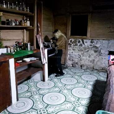 Uma pessoa está cozinhando em uma cozinha rústica com piso de ladrilhos verdes e brancos estampados, prateleiras de madeira, parede de pedra e vários itens de cozinha bem organizados.