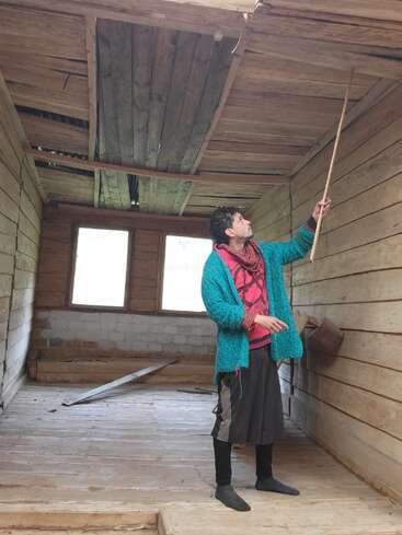 Uma pessoa está descalça em uma sala de madeira, apontando para um teto danificado com uma vara. A sala parece velha, inacabada e precisa de reparos.