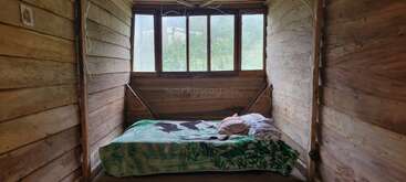 Um quarto simples de madeira apresenta uma cama de solteiro com roupas de cama e travesseiros coloridos, janelas grandes que fornecem luz natural e uma vista externa da vegetação, criando uma atmosfera rústica.