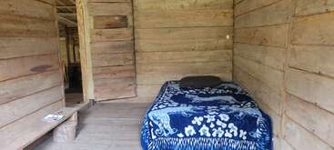 Esta imagem mostra um quarto simples de madeira com paredes de tábuas inacabadas, uma cama coberta com um cobertor azul com estampa de animais, um travesseiro e um pequeno banco.