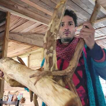 A man inside a wooden cabin holds a large, unfinished tree branch or piece of driftwood, wearing a colorful sweater and looking surprised or focused.