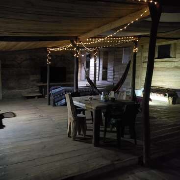Interior de cabana de madeira rústica com luzes de corda, redes, uma mesa de jantar simples, cadeiras de madeira, sofá aconchegante e ambiente descontraído; aconchegante, convidativo e perfeito para reuniões.