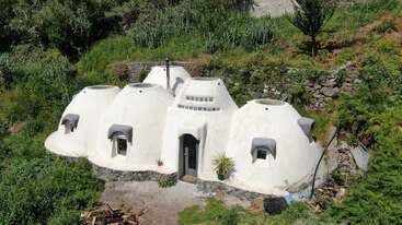 Esta imagen muestra una casa única, con forma de cúpula, construida con sacos de tierra y rodeada de exuberante vegetación, con cuatro cúpulas blancas interconectadas, pequeñas ventanas y una puerta de entrada central.