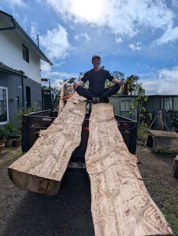 Un hombre está sentado con las piernas cruzadas sobre unos tablones de madera grandes y recién cortados que sobresalen de la caja de un camión, con una casa y un cielo azul al fondo.