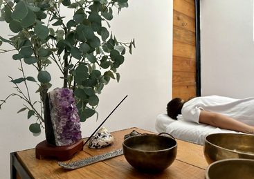 A peaceful spa setting with a person lying face-down on a massage bed, surrounded by crystals, incense, singing bowls, and lush green eucalyptus branches.
