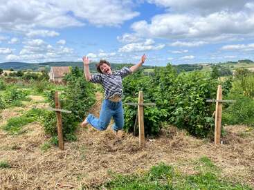Une personne joyeuse saute pieds nus dans un jardin au milieu de plantes vertes luxuriantes, vêtue d'un jean et d'une chemise à rayures. Un ciel lumineux, des nuages gonflés et des collines ondulantes l'entourent.