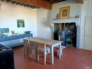 This cozy kitchen features a rustic wooden dining table, four chairs, a large fireplace, wood stove, wall art, sideboard with dishes, and exposed ceiling beams.