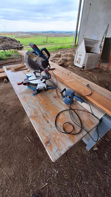A rustic workbench sits outdoors with a miter saw, sander, and wooden planks, surrounded by farmland, tools, and a view of rolling green hills.