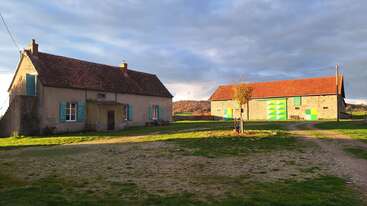 A rural scene with a house and barn, both with red roofs. The house has turquoise shutters, the barn has green doors. Grass and dirt surround them.