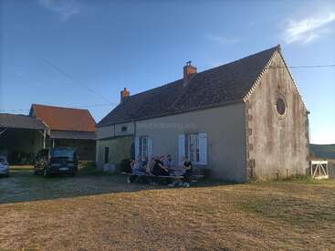 A group of people sit at a picnic table outside a rustic house, enjoying the sunshine. Two cars are parked nearby; the setting feels peaceful.