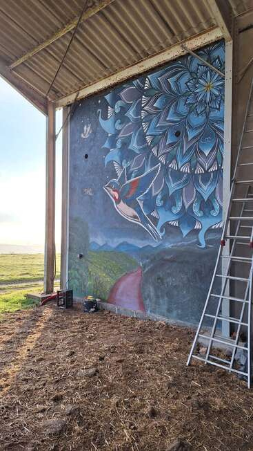Um grande mural na parede mostra um pássaro colorido voando, com um fundo de padrões florais azuis, montanhas e um caminho vermelho. Tem uma escada por perto.