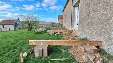 A imagem mostra um canteiro de obras ao lado de uma casa com vigas de madeira colocadas sobre blocos de concreto, preparando as fundações para uma nova estrutura em uma área gramada.