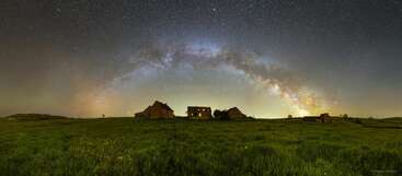 Um céu noturno de tirar o fôlego mostra a Via Láctea em arco sobre casas rústicas em um vasto campo verde, misturando a maravilha cósmica com a beleza rural serena e a tranquilidade.