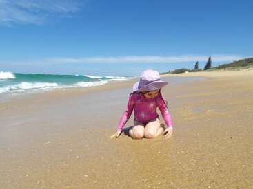 Une jeune fille portant un maillot de bain rose et un chapeau de soleil est agenouillée sur une plage de sable, jouant avec le sable. Le ciel est dégagé et les vagues s'approchent.