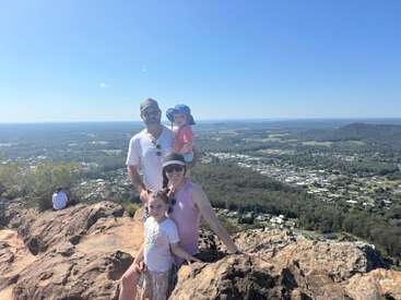 Une famille de quatre personnes pose au sommet d'une montagne rocheuse, souriant sous un soleil radieux. L'arrière-plan pittoresque révèle une ville tentaculaire, des arbres et un horizon lointain.