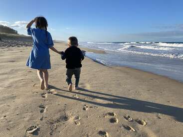 Une fille et un garçon marchent main dans la main sur une plage de sable, projetant de longues ombres, avec les vagues de l'océan et le ciel bleu en arrière-plan. Moment paisible.