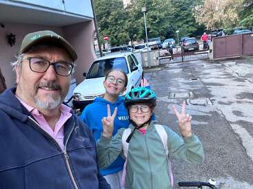 Un homme et deux filles souriantes posent en extérieur. Les filles font le signe de la paix, l'une d'elles porte un casque et un sac à dos, suggérant qu'elles sont prêtes pour une balade à vélo ou pour aller à l'école.