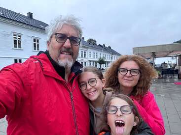 Une famille de quatre personnes, toutes portant des lunettes et des vestes, sourit pour un selfie en extérieur par temps nuageux, avec des bâtiments blancs en arrière-plan. Un enfant tire la langue.