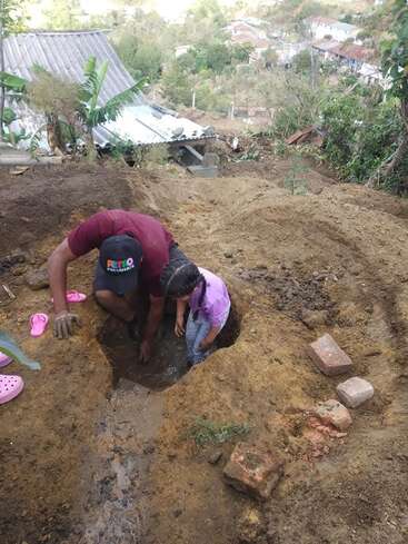 Um homem e uma menina estão cavando um buraco lamacento na encosta de uma colina, com tijolos por perto e sandálias cor-de-rosa espalhadas, o que sugere um ambiente rural.