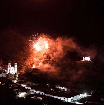 À noite, fogos de artifício iluminam uma pequena cidade com prédios coloniais brancos e igrejas. A fumaça enche o céu, lançando um brilho festivo e dramático sobre a encosta.