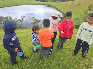 Seis crianças estão juntas em uma colina gramada perto de um lago, vestindo moletons e jaquetas coloridas, aparentemente conversando ou observando algo interessante.