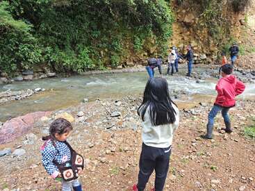Um grupo de crianças e adultos explora a margem rochosa de um rio cercada por uma vegetação exuberante. Alguns atravessam o riacho, enquanto outros observam, curtindo juntos a natureza e a aventura ao ar livre.