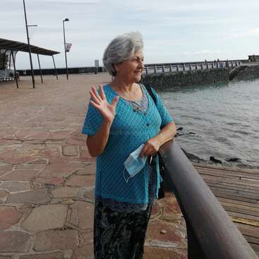 La imagen muestra a una anciana de pie sobre una pasarela de piedra junto a una masa de agua, agitando la mano con una mascarilla quirúrgica azul en la otra.