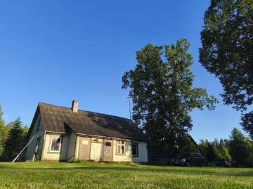 Una rústica casa de madera con tejado oscuro se asienta sobre un frondoso césped verde bajo un cielo azul despejado, rodeada de altos árboles y lejanos coches aparcados.