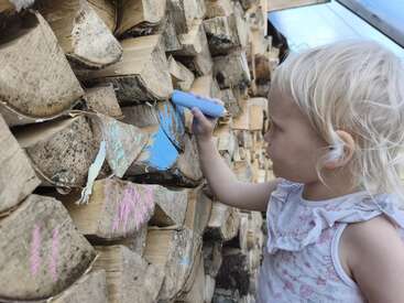 Una niña con el pelo rubio dibuja sobre leña apilada utilizando tiza de colores, añadiendo vibrantes marcas azules y rosas en los troncos de madera rústica al aire libre.