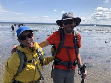 Dos excursionistas sonrientes de pie en un pintoresco llano mareal, con mochilas, gafas de sol y equipo para actividades al aire libre. Un grupo de personas camina al fondo bajo un cielo luminoso.