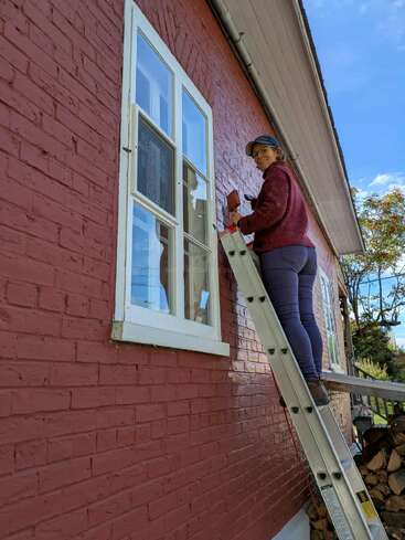 Una persona en una escalera, con sombrero y gafas, pinta el marco de la ventana de una casa de ladrillo rojo en el exterior en un día soleado con cielo azul.