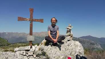 Un hombre está sentado con las piernas cruzadas en la cima rocosa de una montaña, junto a una cruz de madera y un montículo de piedras, disfrutando del cielo despejado y de las pintorescas colinas lejanas.
