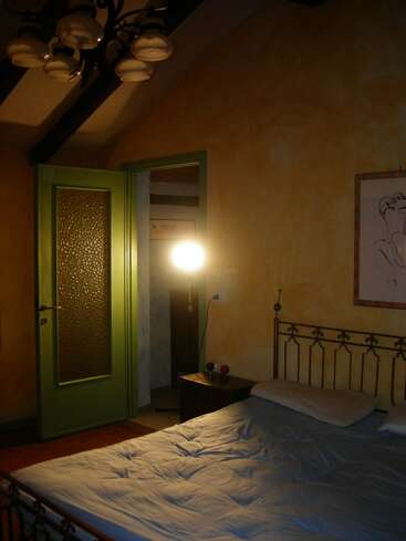 Esta imagen muestra un dormitorio con poca luz, con una cama de hierro forjado, ropa de cama arrugada, una puerta verde, una mesita de noche, una lámpara encendida, paredes amarillas y obras de arte. Ambiente acogedor.
