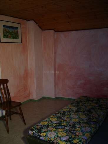 Esta imagen muestra una habitación sencilla y poco iluminada con paredes de textura rosa, una silla de madera, un colchón con motivos florales en el suelo y un cuadro con un marco de colores.