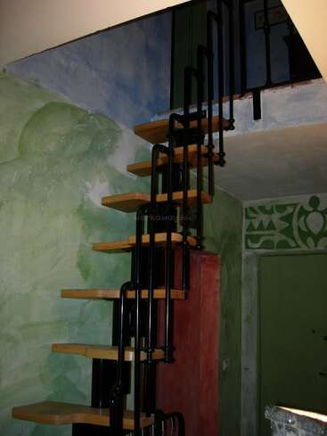 Una estrecha escalera de caracol con peldaños de madera y barandillas de metal negro asciende a la planta superior. Las paredes están pintadas con diseños abstractos en verde, azul y rojo.