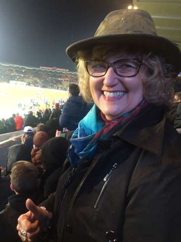 L'image montre une femme portant un chapeau et un manteau lors d'un événement sportif, avec une foule de personnes à l'arrière-plan et un terrain avec des joueurs.