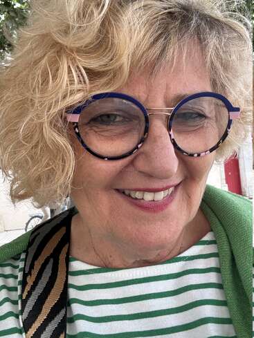 Une femme souriante aux cheveux blonds bouclés porte de grandes lunettes rondes bleues et un t-shirt rayé vert et blanc ; elle se tient debout à l'extérieur, avec un vélo en arrière-plan.