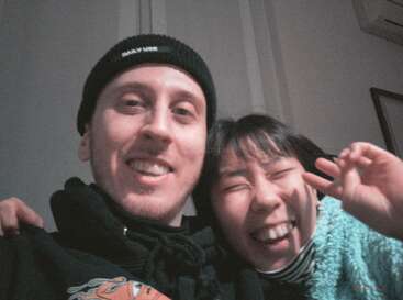 A joyful selfie of two friends, one wearing a beanie and a hoodie, the other smiling widely and making a peace sign, indoors with light walls.