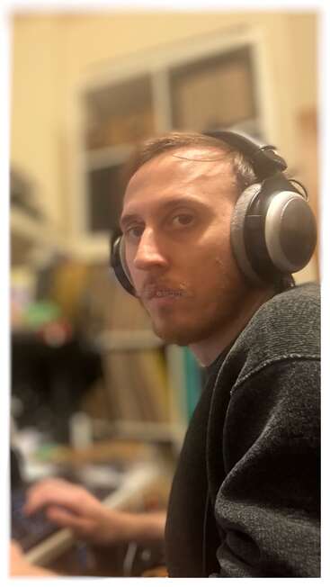A man with light brown hair and beard wears large headphones and a dark sweater. He sits at a desk, looking intently toward the camera, indoors.