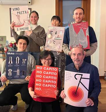 Six people pose indoors, smiling and holding colorful posters with various designs and text. The atmosphere appears friendly and creative, suggesting a casual gathering or celebration.