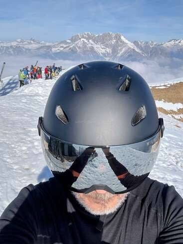 A person wearing a black ski helmet and reflective goggles takes a selfie on a snowy mountain, with other skiers and majestic peaks in the background.