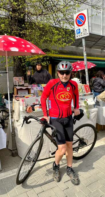 A cyclist in red gear poses with his bike in front of a street market. A woman behind the table makes a peace sign, bright umbrellas overhead.