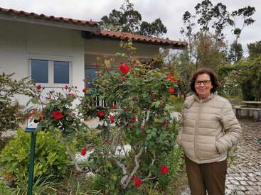 The image depicts a woman standing in front of a rose bush, wearing a beige puffer jacket and brown pants, with a house and trees visible in the background.