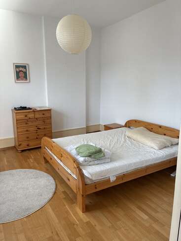 Um quarto simples e arrumado com uma cama de madeira, cômoda e mesa de cabeceira. As paredes brancas, o tapete redondo, a lanterna de papel e o retrato emoldurado criam uma atmosfera aconchegante.