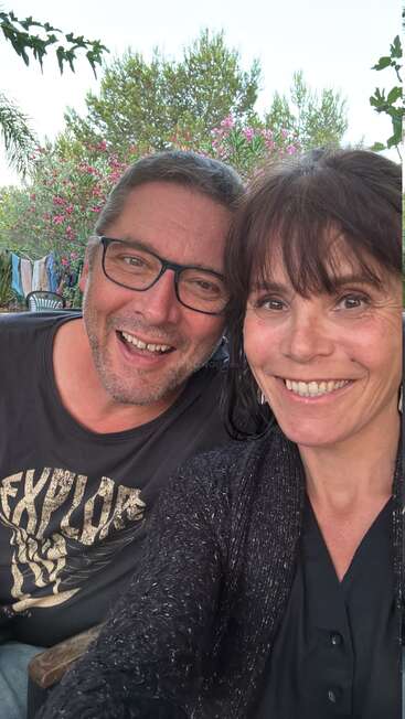 Um homem e uma mulher sorridentes tiram uma selfie ao ar livre. Atrás deles, há árvores verdes, flores cor-de-rosa e roupas penduradas, criando uma atmosfera alegre e descontraída.