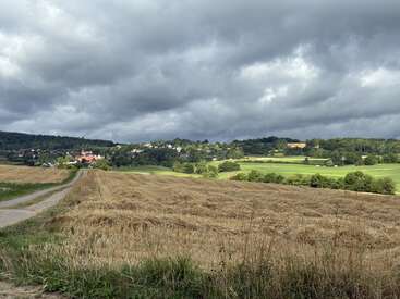 Uma paisagem rural com campos colhidos, um caminho sinuoso, colinas verdejantes e uma pequena aldeia à distância, sob um céu nublado e dramático. Campo tranquilo.