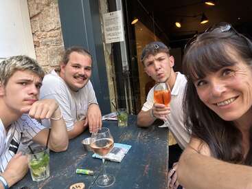 Quatro pessoas se sentam ao redor de uma mesa ao ar livre, tomando bebidas e sorrindo. O ambiente é descontraído e amigável, com um fundo rústico e uma atmosfera aconchegante e acolhedora.