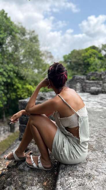 Una mujer con mechas rosas en el pelo está sentada sobre unas antiguas ruinas de piedra, contemplando pensativa el verde bosque, vestida con un vestido ligero y sandalias bajo un cielo nublado.
